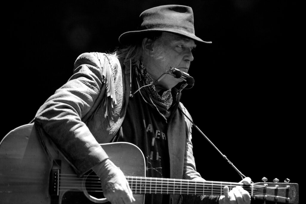 Musician playing guitar with harmonica and hat.