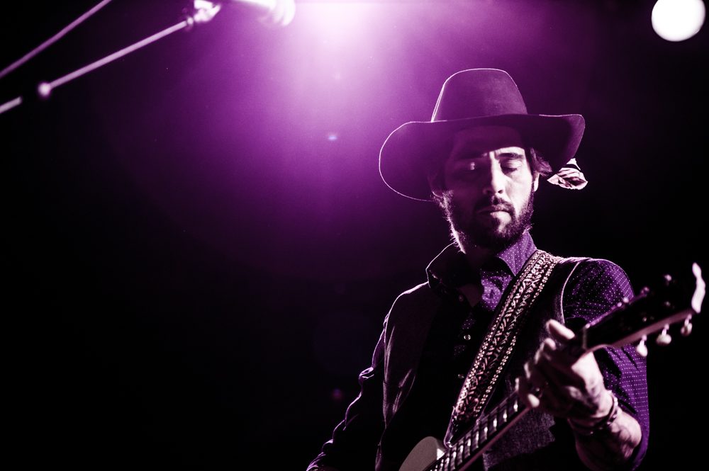 Guitarist performing on stage under purple light.