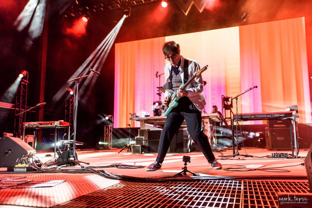 Guitarist performing energetically on stage with lights.