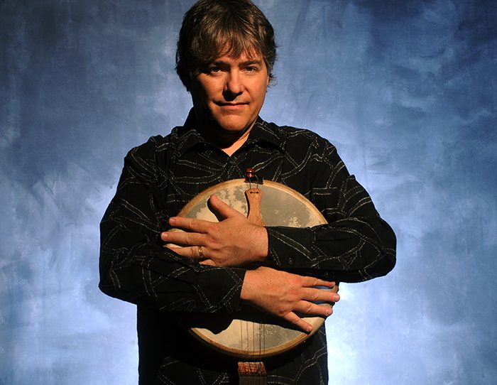 Man holding a banjo against blue backdrop.