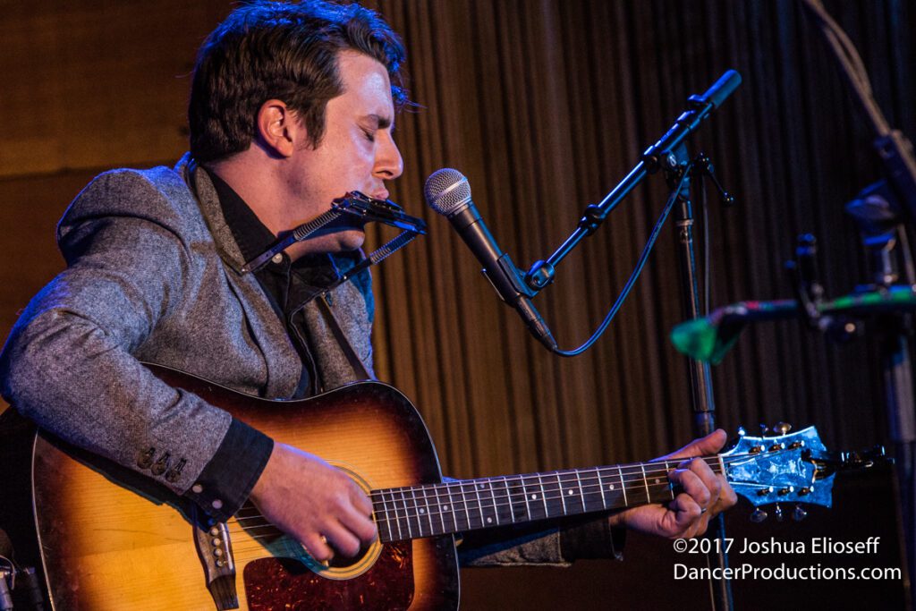 Musician playing guitar and harmonica on stage.