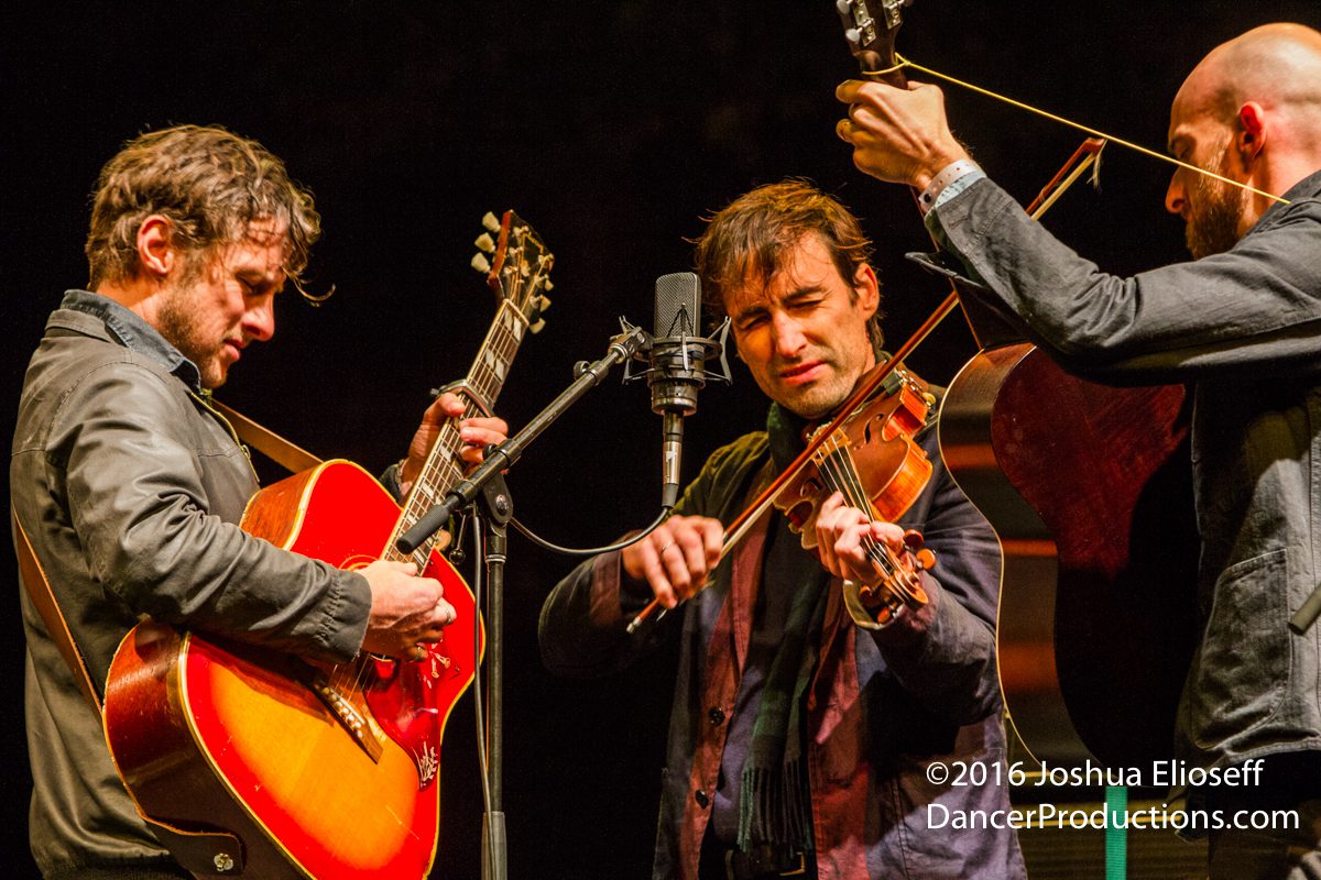 Three musicians performing with guitars and violin.