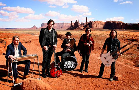 Band posing in desert with musical instruments.