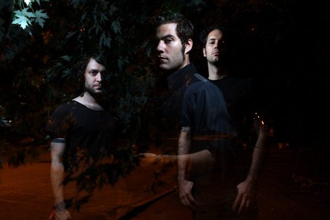 Three men standing in dark, leafy surroundings.