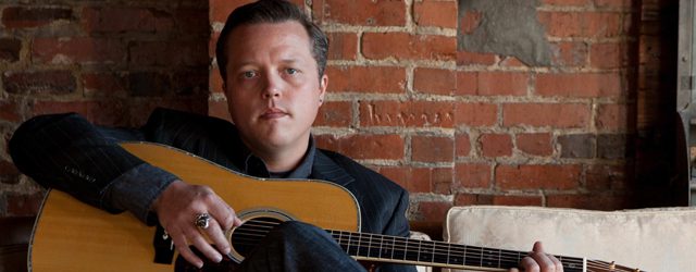 Man sitting with guitar against brick wall.