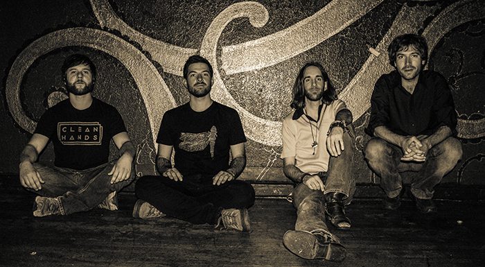 Four men sitting against a decorative wall.