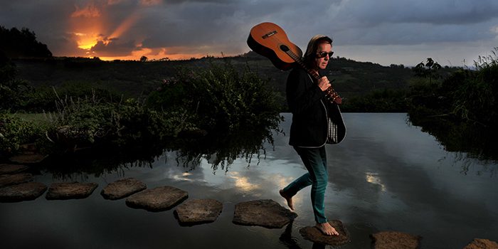 Person crossing river at sunset with guitar.