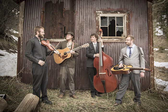 Four musicians playing instruments near rustic building.