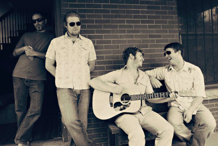 Four men with guitar, sitting and smiling.