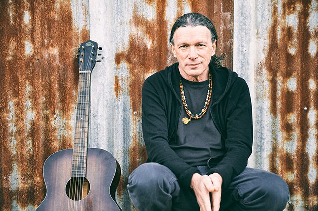 Man sitting with guitar against rustic wall.