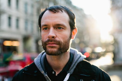 Man standing outdoors, city background, sunlight behind.