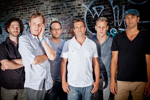 Six men standing against graffiti-covered wall.