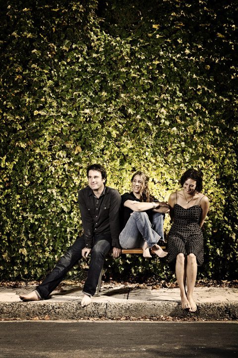 Three people sitting against a leafy wall.