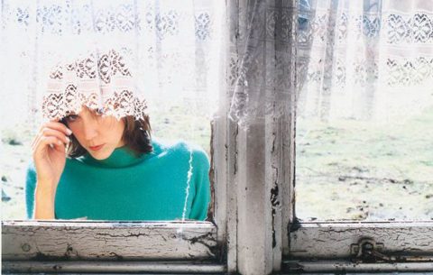Woman peeking through lace curtain by window.