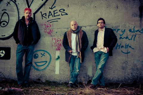Three men standing against graffiti-covered wall.