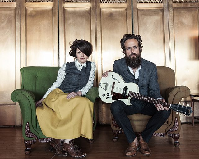 Man with guitar and woman sitting indoors.