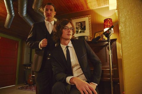 Two men in suits by a piano.