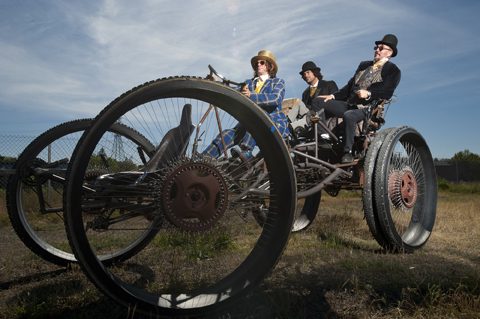 Three people riding a large, vintage tricycle.
