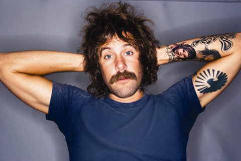 Man with tattoos and mustache, arms raised.
