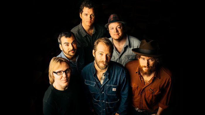Six men posing together, dark background.
