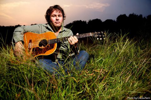 Man playing guitar in grassy field at sunset.