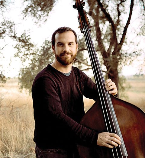 Man playing double bass outdoors near trees.