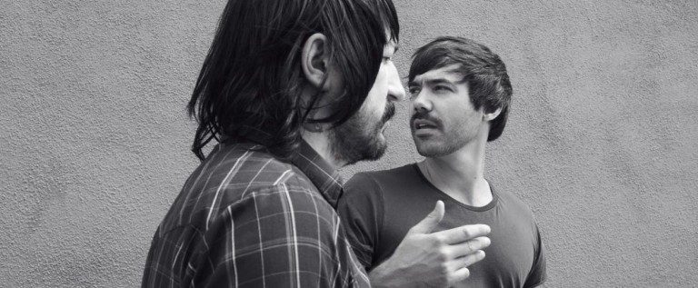Two men talking against a wall.