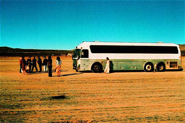 Bus on desert road with people nearby.