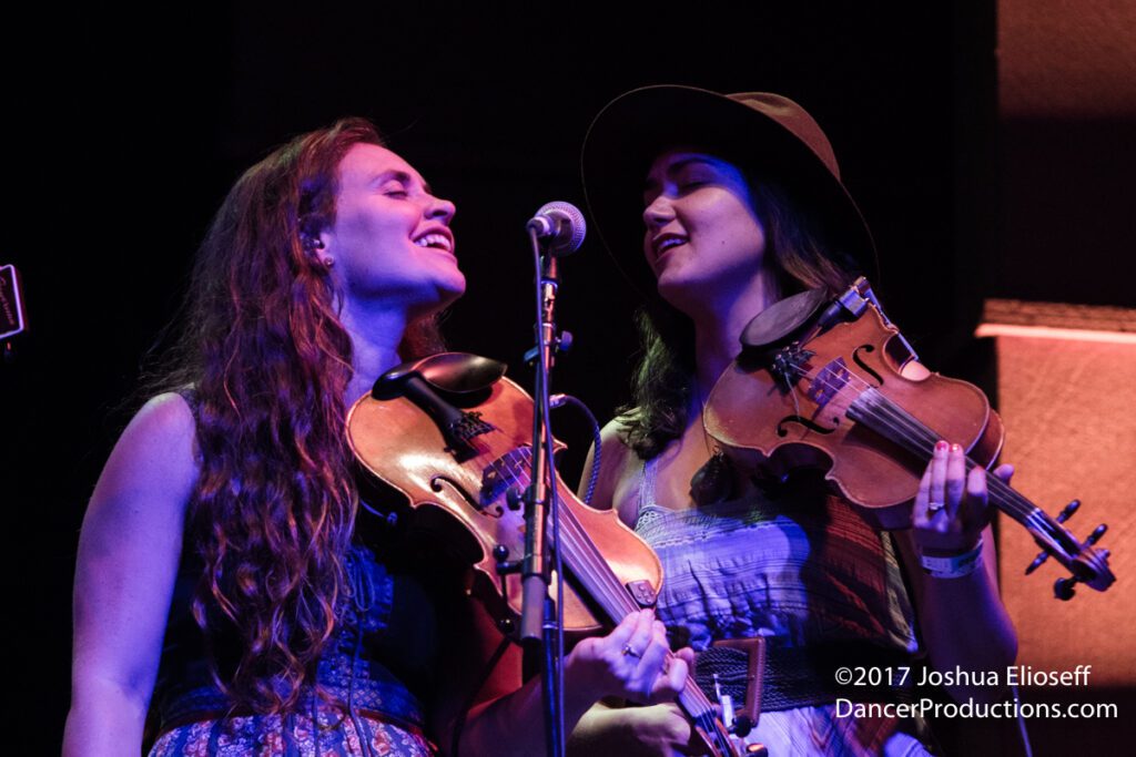 Two women joyfully playing violins on stage.