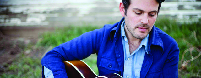 Man playing guitar outdoors, wearing blue jacket.