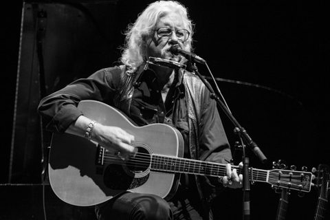 Musician playing guitar and harmonica on stage.