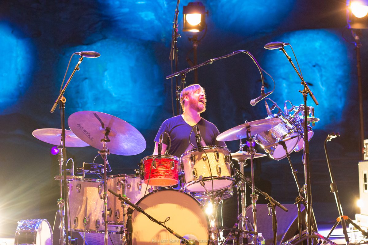 Drummer performing energetically on stage, blue lighting.