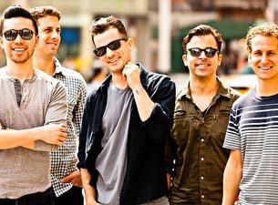 Five smiling men standing outdoors, wearing sunglasses.