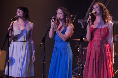 Three women singing on stage with microphones.
