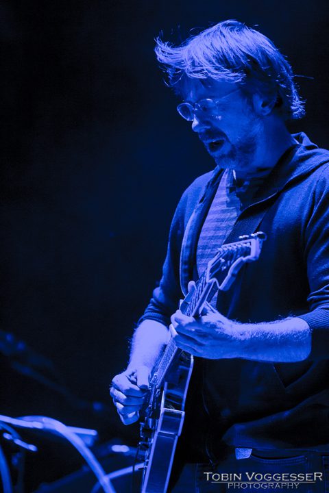 Musician playing guitar on stage, blue lighting.