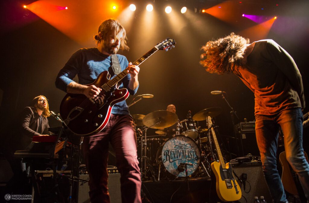 Band performing energetically on stage with lights.