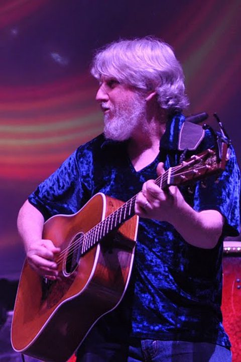 Guitarist performing on stage with colorful backdrop.