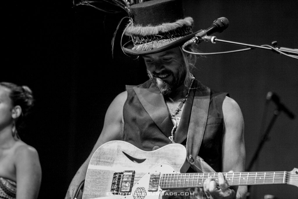 Musician playing guitar on stage, wearing hat.