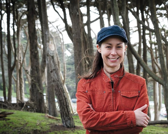 Woman smiling in forest, wearing orange jacket.