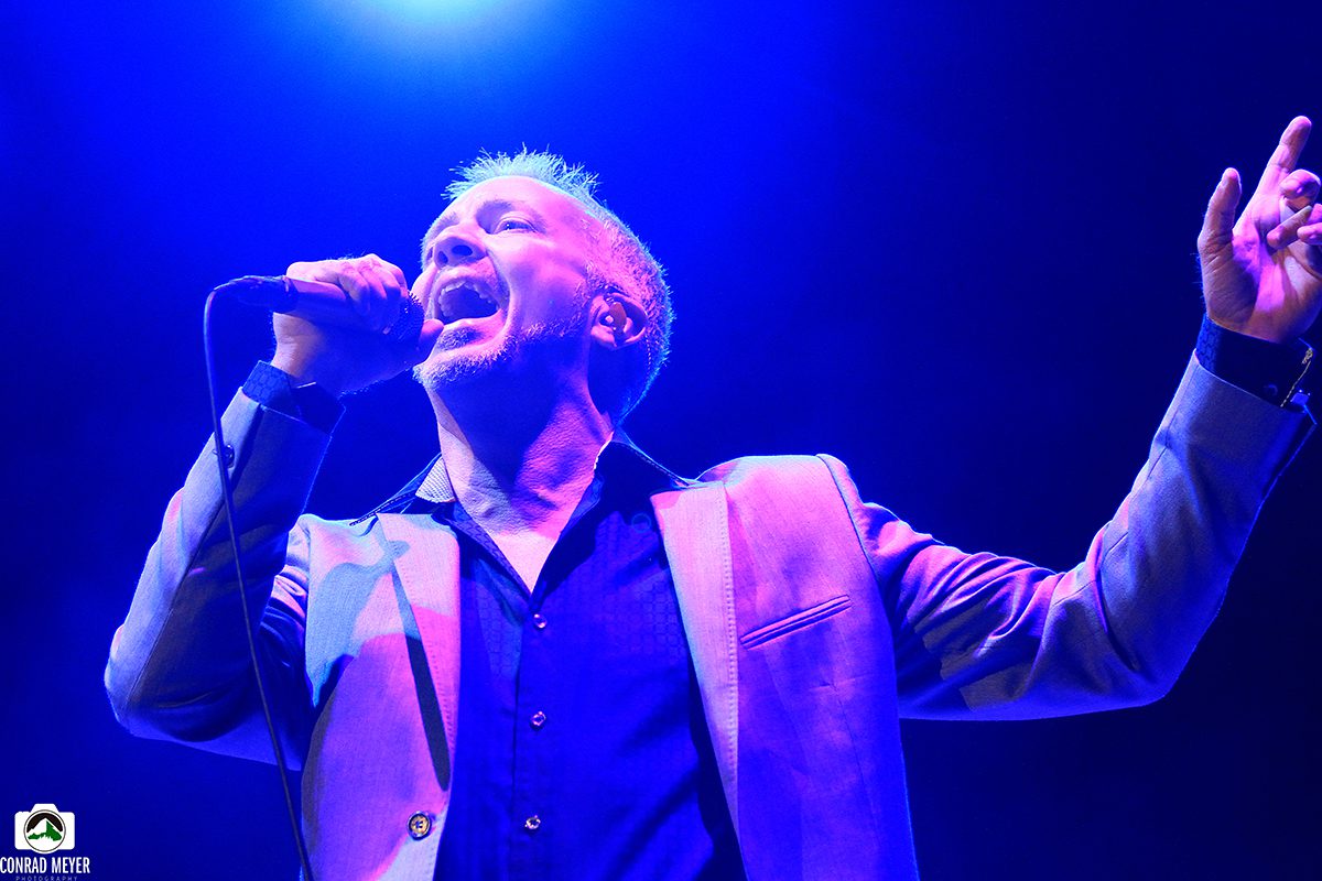 Singer performing energetically on stage, blue lighting.