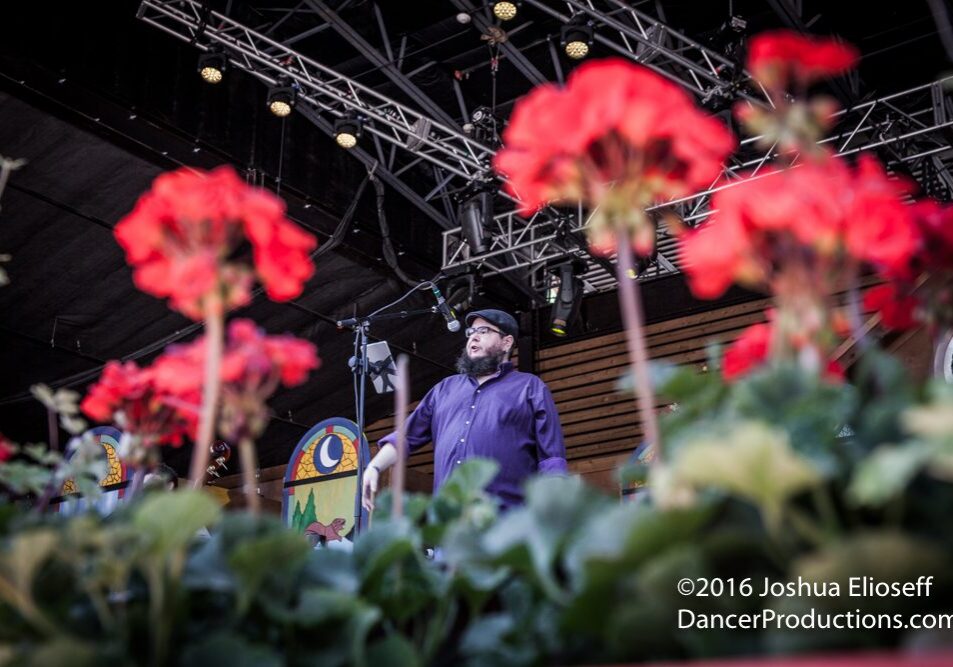Man on stage with red flowers foreground.