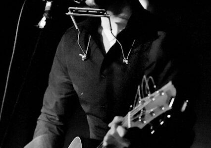 Musician performing with guitar and harmonica.