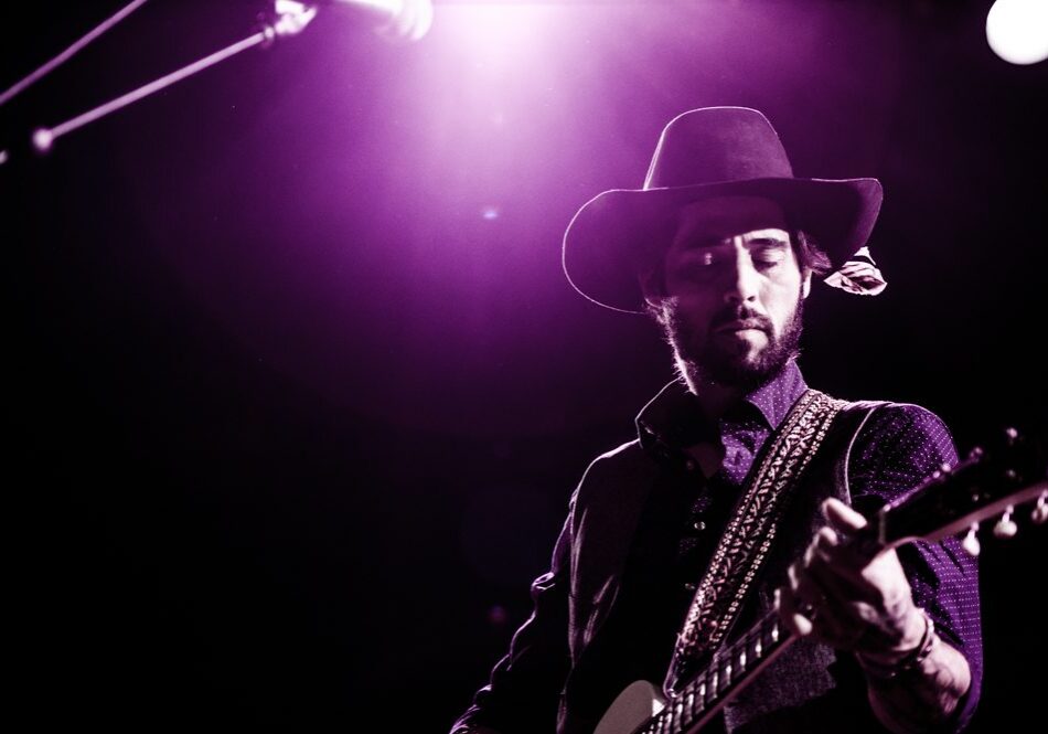 Guitarist performing on stage under purple light.