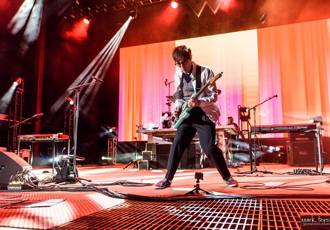 Guitarist performing energetically on stage with lights.
