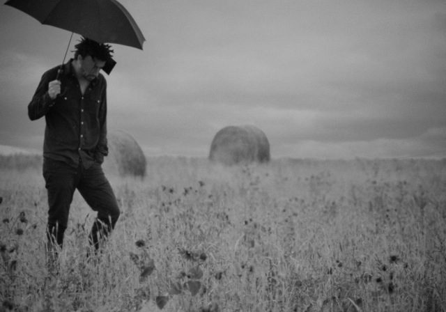 Man with umbrella in field, gray sky.