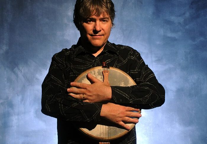 Man holding a banjo against blue backdrop.