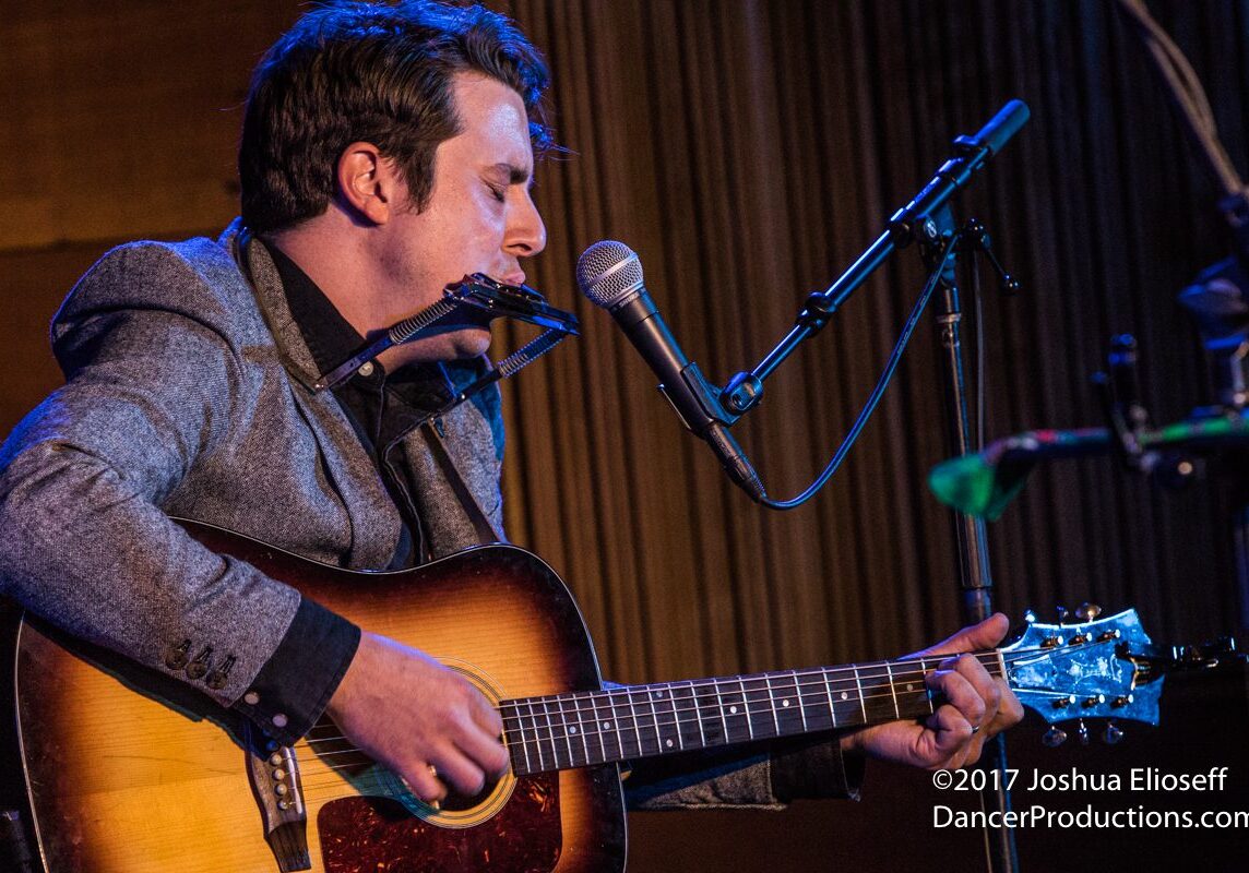 Musician playing guitar and harmonica on stage.