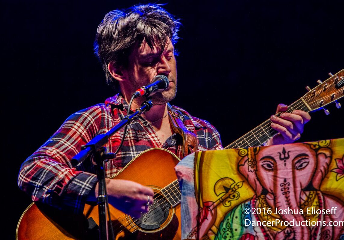 Musician playing guitar on stage, colorful backdrop.