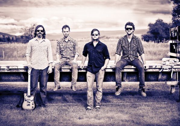 Four men posing outdoors with guitar.