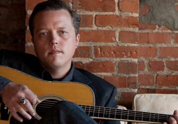 Man sitting with guitar against brick wall.